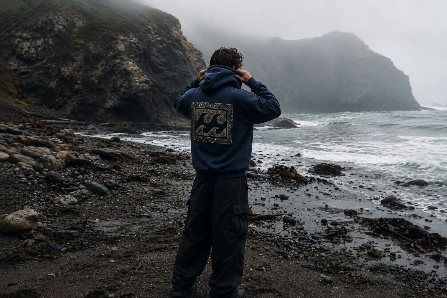 Person in a Driftline hoodie facing a cold, rocky coastline